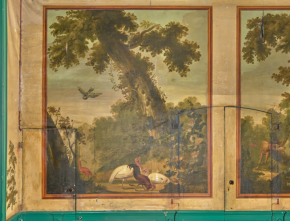 Detail van de beschilderde wand in de eetzaal: een rode ibis onder een grote boom, met gevogelte op de voorgrond