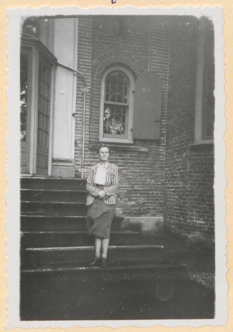 Zwart-witportret van een vrouw met opgestoken haar in een gestreept jasje en knielange rok, staand met gevouwen handen op de stenen buitentrap van het kasteel
